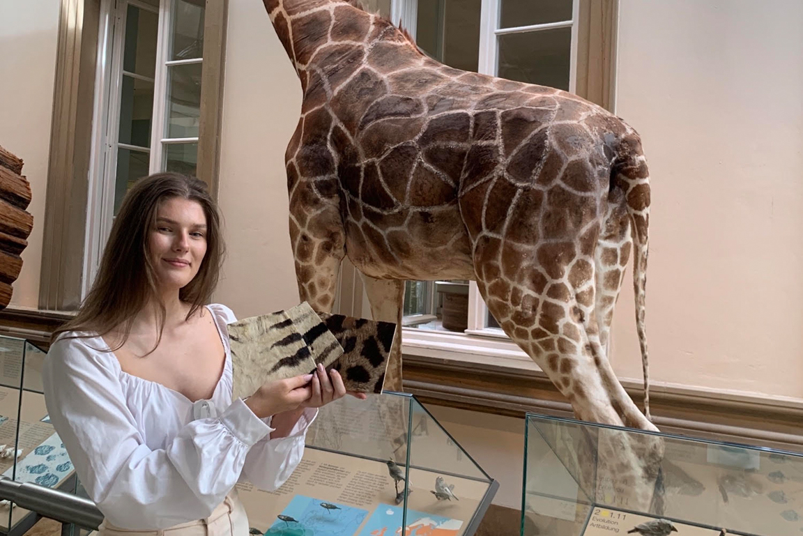Junge Frau steht vor einer präparierten Giraffe und hält Fellstücke verschiedener Tiere.