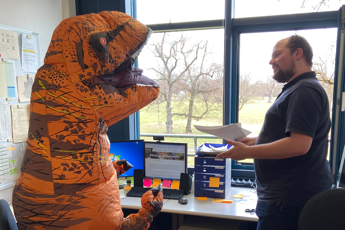 Ein junger Mann mit Dokumenten in der Hand redet mit einer Person, die ein aufblasbares, orangenes Dinokostüm trägt.